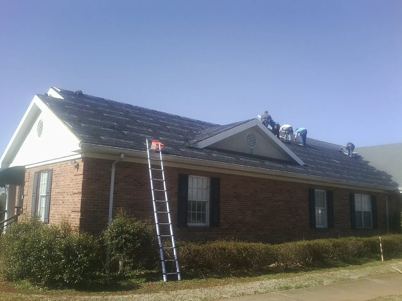 Crew tearing shingles off a brick ranch roof with a ladder set — Emergency Roof Repair work by our crew in Ouaquaga