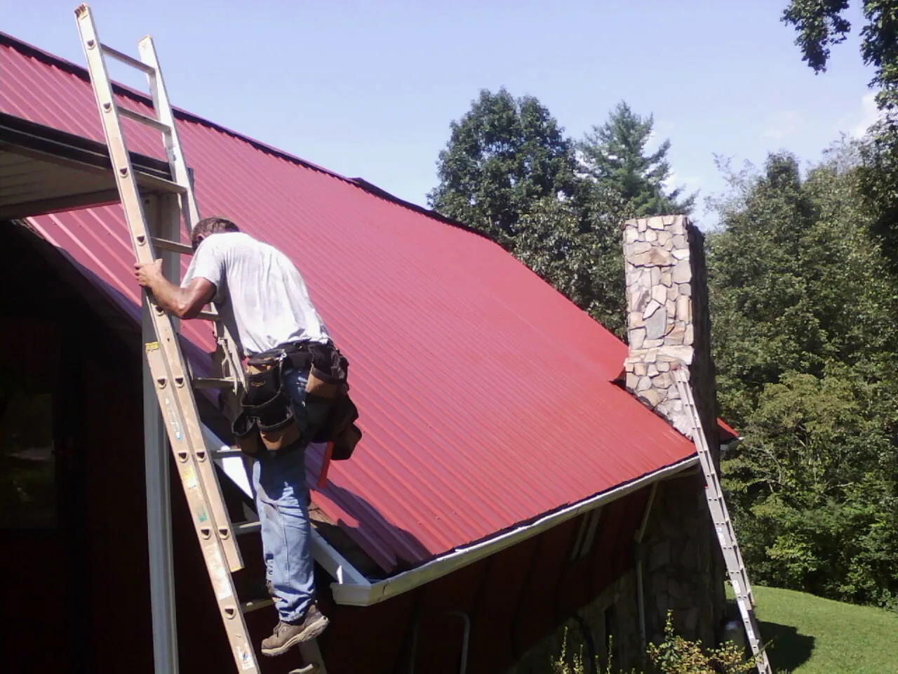 Roofer on a ladder beside a red metal roof and stone chimney — Soffit & Fascia Repair by Roofing Brothers in Ouaquaga