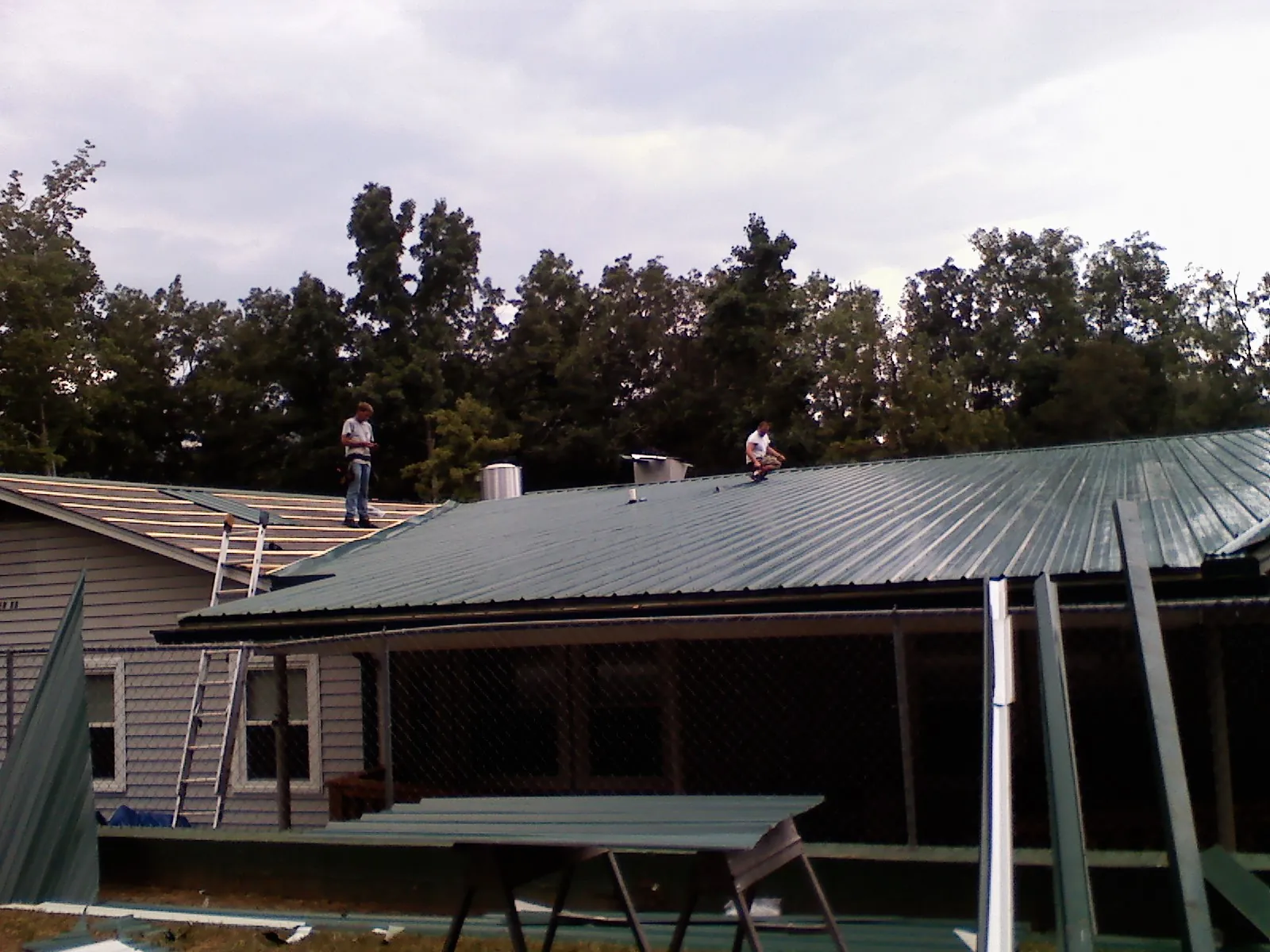 Two roofers installing green metal panels above a covered porch in Ouaquaga